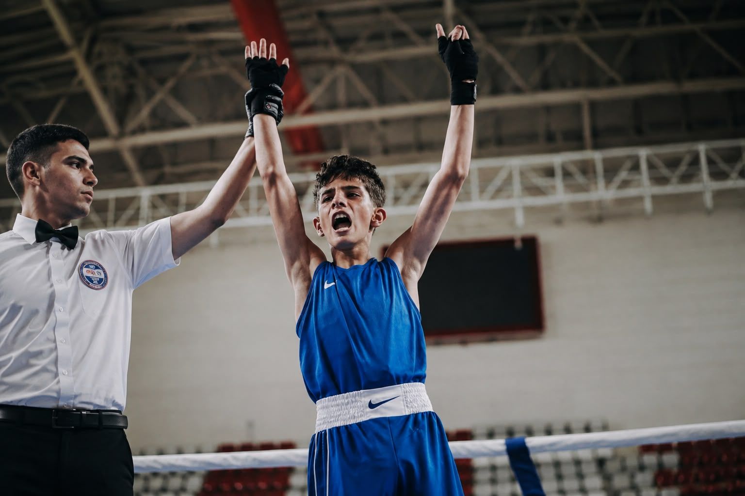 Boks ringinde mavi forması ve eldivenleriyle genç sporcu, bir hakemin kaldırdığı eliyle kollarını havaya açıyor.