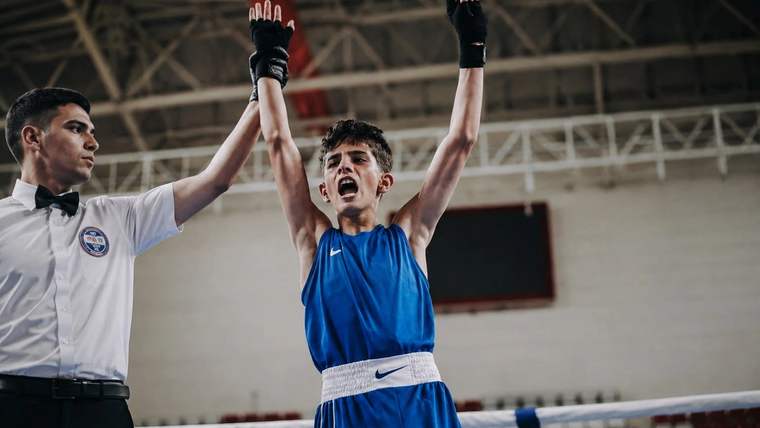Boks ringinde mavi forması ve eldivenleriyle genç sporcu, bir hakemin kaldırdığı eliyle kollarını havaya açıyor.