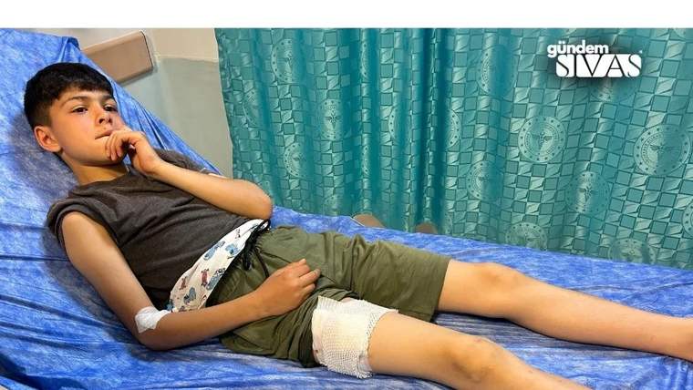 Yatakta uzanan bir çocuğun kolunda ve dizinde bandaj, arka planda mavi örtü ve desenli perde görülüyor.