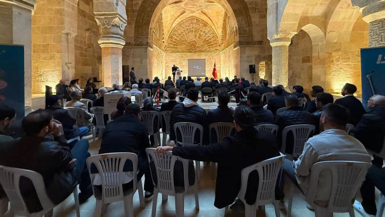 Taş kemerli bir salonda sandalyelerde oturan kalabalık topluluk ve önde sunum yapılan kürsü görülüyor.