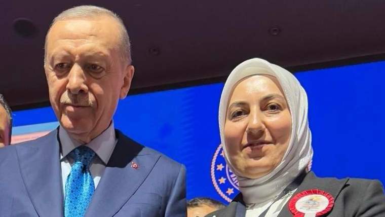 Bir kadın, yanında bir erkekle birlikte sahnede duruyor; kadının üzerinde rozet var.