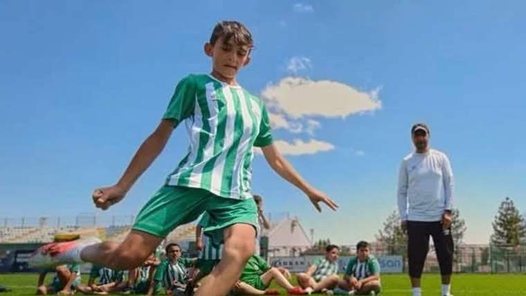 Yeşil-beyaz formasıyla bir çocuk futbol topuna vuruyor, arka planda diğer çocuklar ve bir antrenör var.