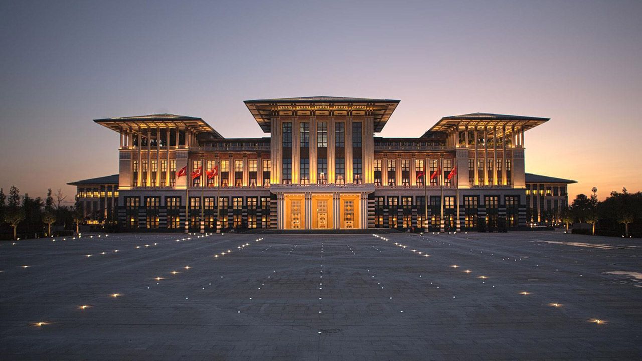 Geniş bir meydanın karşısında, akşam ışıklarıyla aydınlatılmış simetrik ve görkemli bir yapı görülüyor.