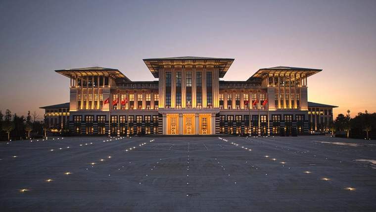 Geniş bir meydanın karşısında, akşam ışıklarıyla aydınlatılmış simetrik ve görkemli bir yapı görülüyor.
