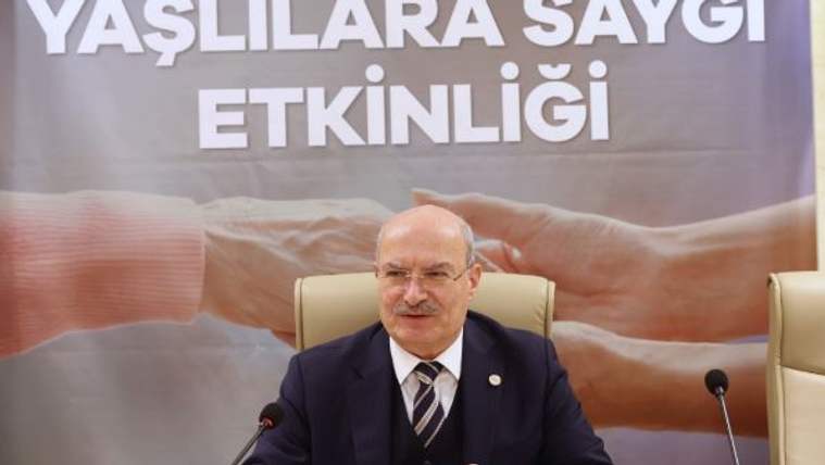 “Yaşlılara Saygı Etkinliği” yazılı pankart önünde, masada oturan takım elbiseli bir kişi