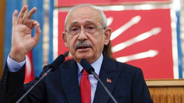 Kürsüde iki mikrofon önünde konuşan, kırmızı kravatlı bir kişi elini kaldırarak işaret yapıyor.