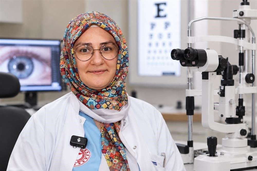 Renkli başörtülü ve gözlüklü bir kişi, göz muayene cihazının yanında kameraya bakıyor.