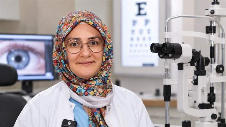 Renkli başörtülü ve gözlüklü bir kişi, göz muayene cihazının yanında kameraya bakıyor.