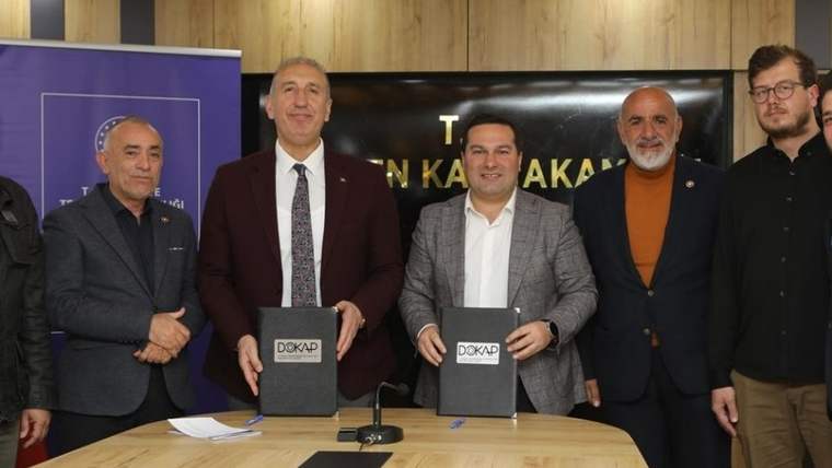 Masada oturan dört kişi, ellerindeki DÖKAP yazılı dosyaları kameraya doğru tutuyor; arkada kürsü bulunuyor.