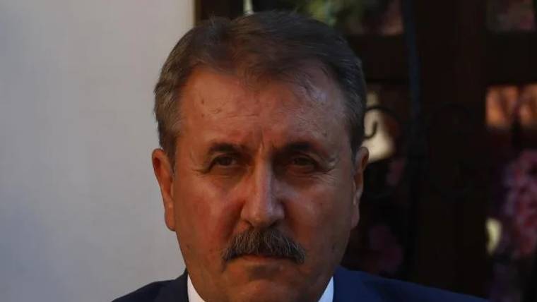 Koyu mavi takım elbise giymiş, kısa saçlı ve bıyıklı bir adamın yüzü, arka planda bulanık bir ortam var.