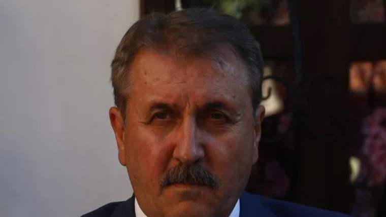 Koyu mavi takım elbise giymiş, kısa saçlı ve bıyıklı bir adamın yüzü, arka planda bulanık bir ortam var.
