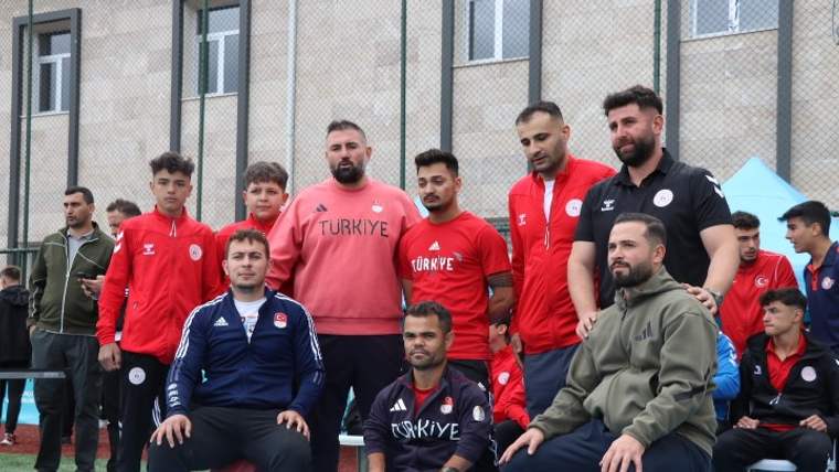 Spor sahasında kırmızı formalar giyen halterciler ve antrenörler, kamera karşısında birlikte duruyor.