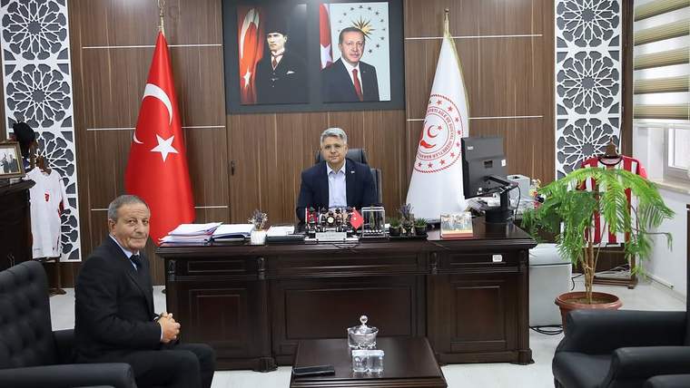Bir ofiste, masanın arkasında oturan bir kişi ve önünde oturan başka bir kişi görünüyor. Duvarlarda Türk bayrağı ve Cumhurbaşkanı'nın resmi var.