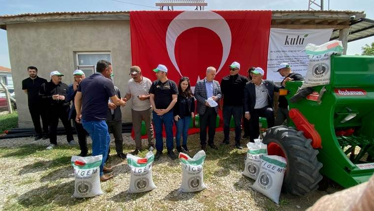 Kırmızı-beyaz Türk bayrağının arkasında, kenevir ekimi için toplanmış bir grup insan ve yanlarında çuvallar var.