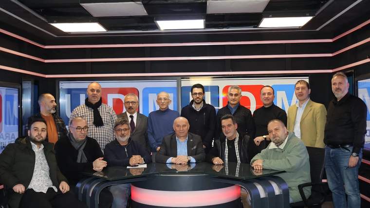 Televizyon stüdyosunda, yuvarlak masanın çevresinde oturan ve ayakta duran bir grup kişi görülüyor.