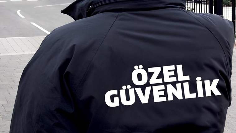 Sırtında “ÖZEL GÜVENLİK” yazan koyu renkli montlu bir kişi, kaldırım kenarında duruyor.