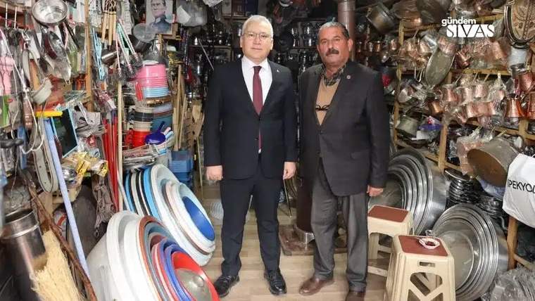 Hırdavat dükkânında takım elbiseli iki kişi, çeşitli metal ürünler ve temizlik gereçleri arasında duruyor.