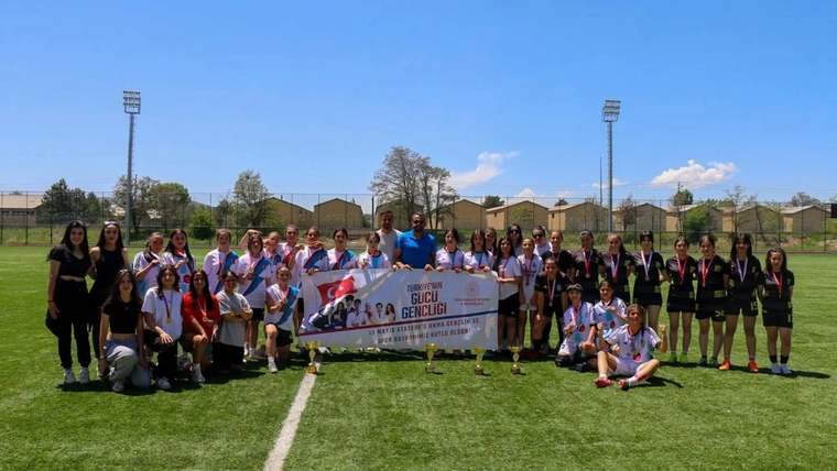 Futbol sahasında, ellerinde Türk bayrağı ve pankart tutan gençler ile sporcular toplu halde duruyor.