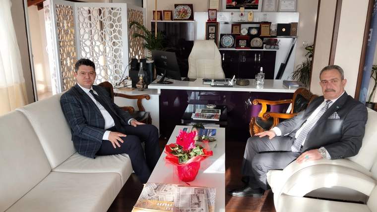 Ofiste koltuklarda oturan iki erkek, aralarındaki masada kırmızı çiçek buketi ve çeşitli yayınlarla görülüyor.