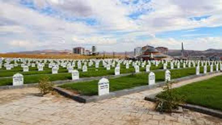 Beyaz mezar taşlarının sıralandığı bir mezarlık alanı, arka planda binalar ve bulutlu bir gökyüzü görünmekte.