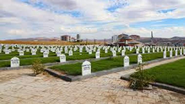 Beyaz mezar taşlarının sıralandığı bir mezarlık alanı, arka planda binalar ve bulutlu bir gökyüzü var.