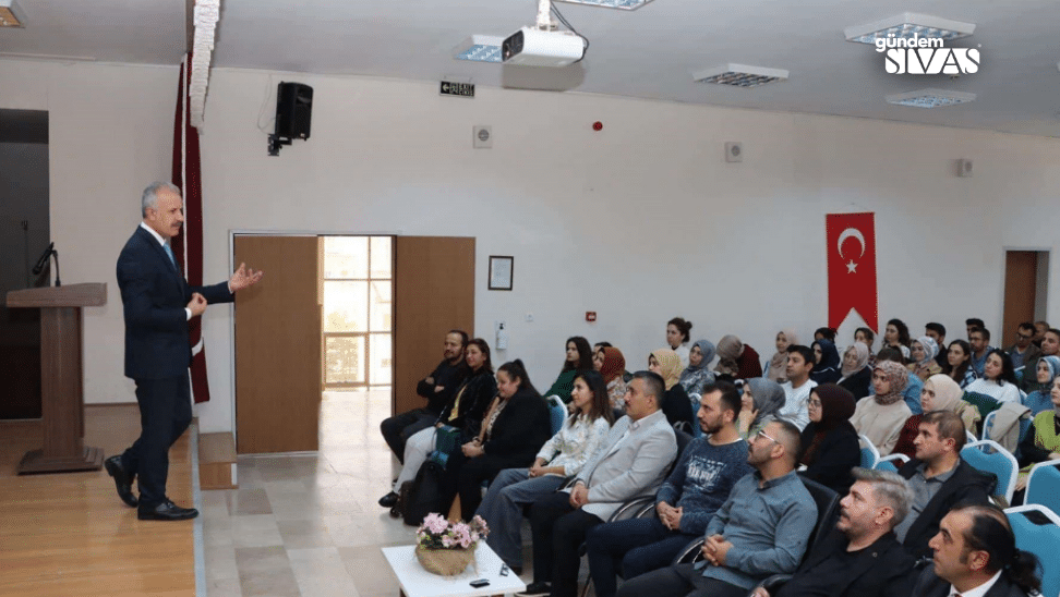 Bir salonda, sahnede konuşan bir kişi ve dinleyici olarak oturan kalabalık.