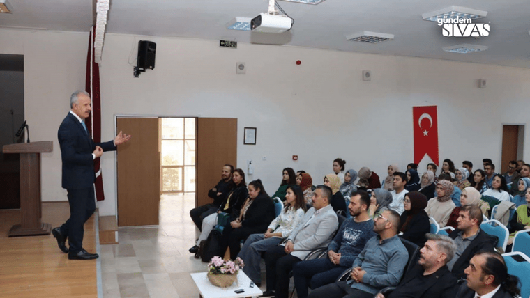 Bir salonda, sahnede konuşan bir kişi ve dinleyici olarak oturan kalabalık.