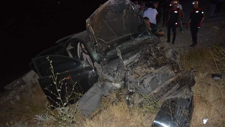 Gece karanlığında ağır hasar almış otomobilin yanında üç görevli ve çevrede kuru otlar görülüyor.