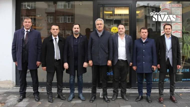 Bir grup erkek, bir dükkânın önünde sırada duruyor. Arkada dükkânın camında yazılar var.