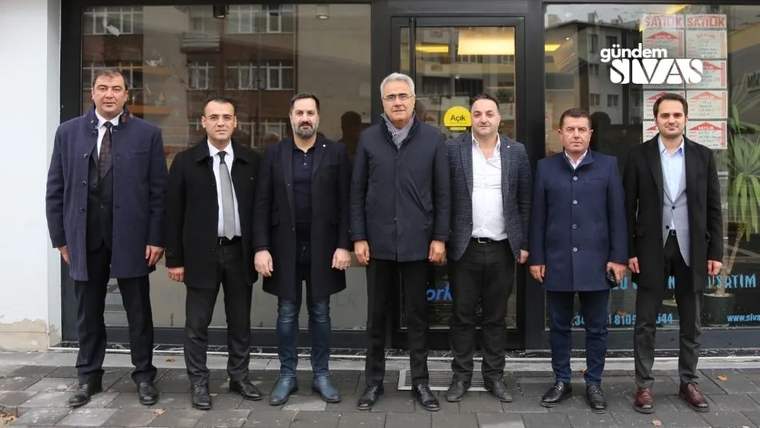 Bir grup erkek, bir dükkânın önünde sırada duruyor. Arkada dükkânın camında yazılar var.