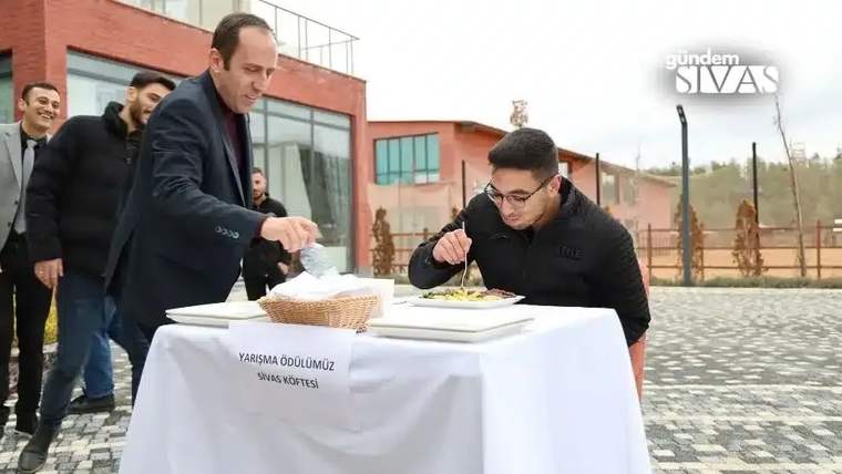 Açık havada masadaki tabaktan yemek yiyen bir kişi ve yanında duran başka bir kişi görülüyor.