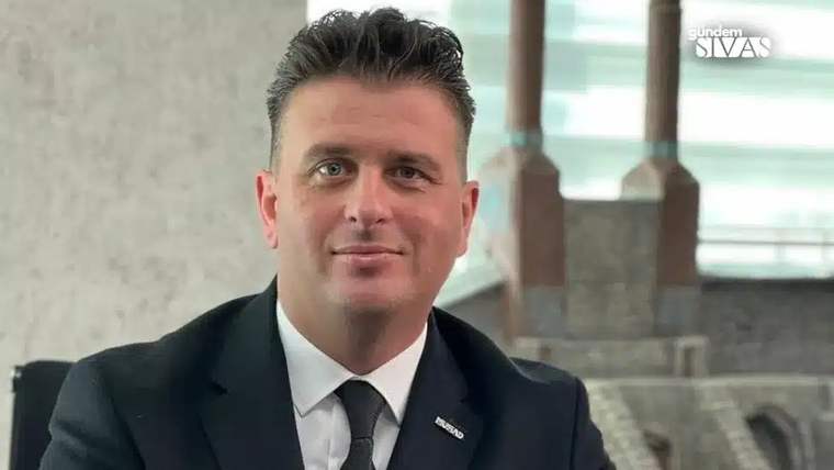 Koyu takım elbise giymiş bir kişi, gülümseyerek kameraya bakıyor.
