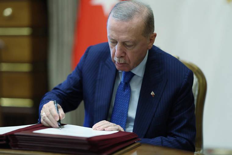 Koyu takım elbiseli bir kişi, masadaki belgeleri kalemle imzalarken görülüyor.