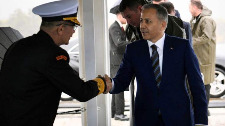 Bir kişi, deniz kıyısında üniformalı bir kişiyle tokalaşıyor.