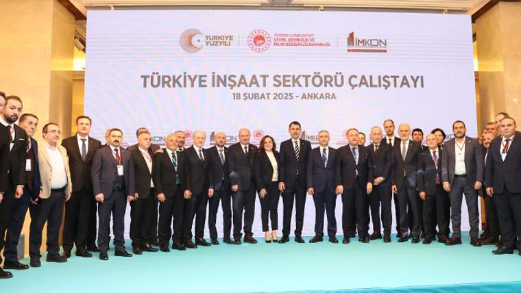 Çalıştay afişi önünde takım elbiseli kalabalık bir grup, sahnede yan yana duruyor.