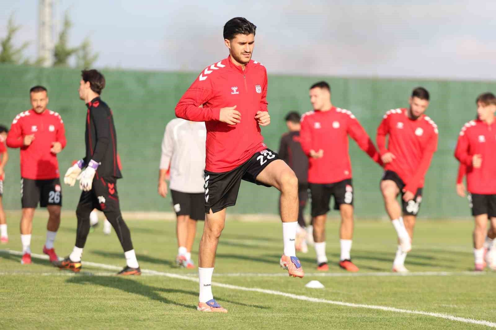 Kırmızı formalı futbolcular çim sahada antrenman yaparken öndeki oyuncu dizini kaldırıyor.