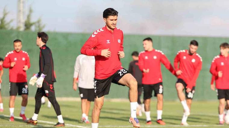 Kırmızı formalı futbolcular çim sahada antrenman yaparken öndeki oyuncu dizini kaldırıyor.