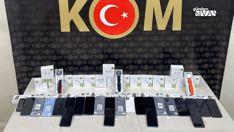 Bir masanın üzerinde çeşitli elektronik cihazlar, kutular ve telefonlar sergileniyor.