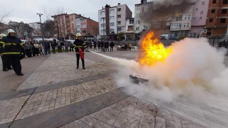 Kent meydanında itfaiyeciler ve izleyenler önünde alev alan düzeneğe su ve köpük püskürtülüyor.