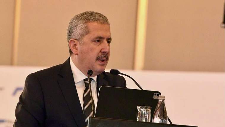 Takım elbiseli, bıyıklı bir kişi kürsüde mikrofon ve dizüstü bilgisayarın önünde konuşuyor.