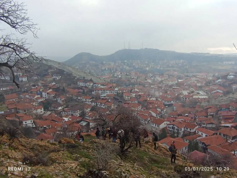 Kayalık bir yamaçta yürüyen insanlar, kırmızı çatılı evlerin ve tepelerin bulunduğu yerleşimi izliyor.