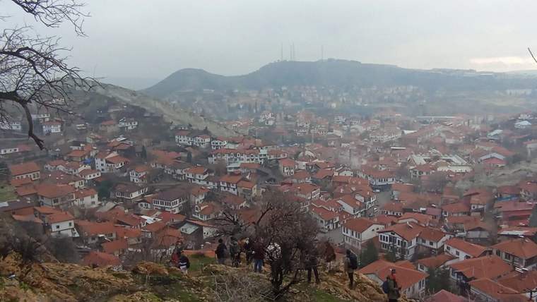 Kayalık bir yamaçta yürüyen insanlar, kırmızı çatılı evlerin ve tepelerin bulunduğu yerleşimi izliyor.