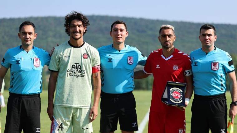 Çim sahada üç hakem ve iki futbolcu yan yana duruyor; kırmızı formalı futbolcu elinde plaket tutuyor.