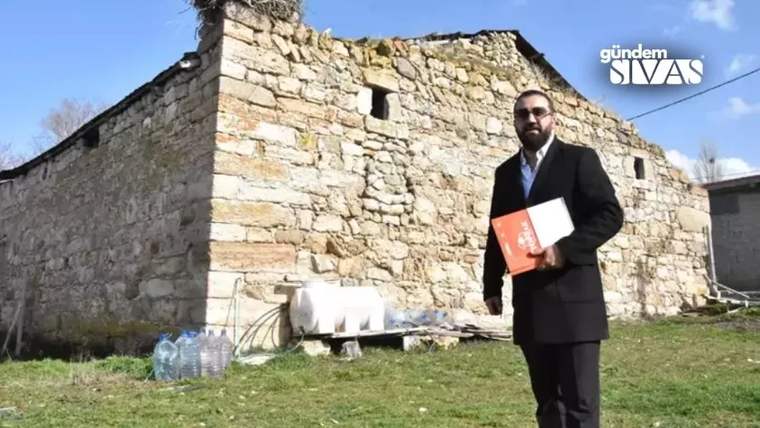 Taş duvarlı eski bir yapının önünde, elinde kırmızı dosya tutan takım elbiseli bir kişi duruyor.