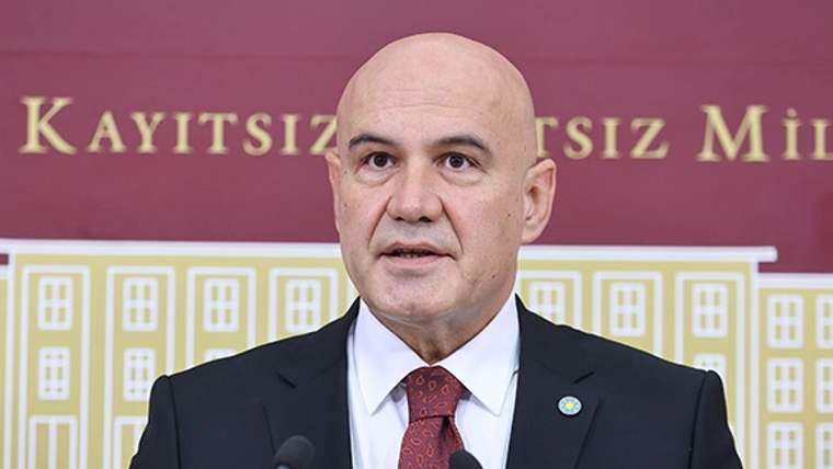 Takım elbiseli bir kişi, iki mikrofonun önünde konuşurken arkasında yazılı bir pano görülüyor.