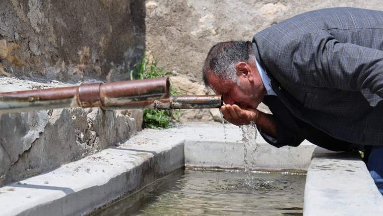 Bir adam, taş bir çeşmeden su içiyor. Çeşmenin etrafında taş duvarlar var.