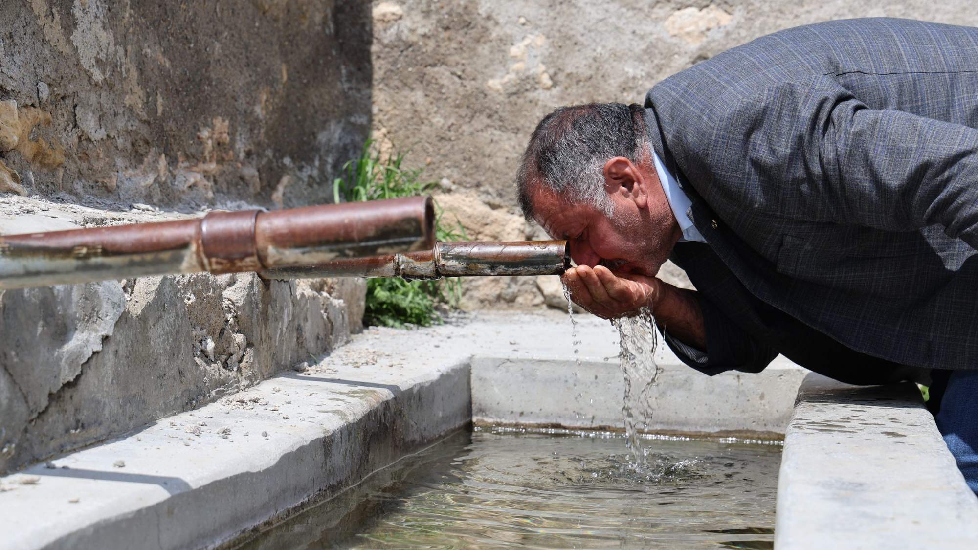 Bir adam, taş bir çeşmeden su içiyor. Çeşmenin etrafında taş duvarlar var.