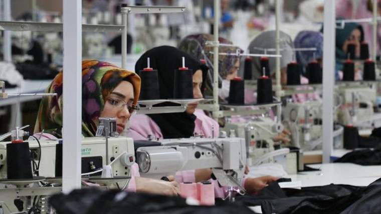 Tekstil atölyesinde üç kadın, dikiş makinelerinde kumaş parçaları üzerinde çalışıyor; çevrede iplik makaraları bulunuyor.
