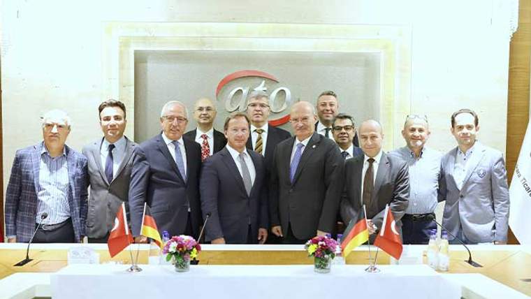 Toplantı salonunda ATO logosu önünde duran kalabalık grup, masadaki Türk ve Alman bayraklarıyla görülüyor.