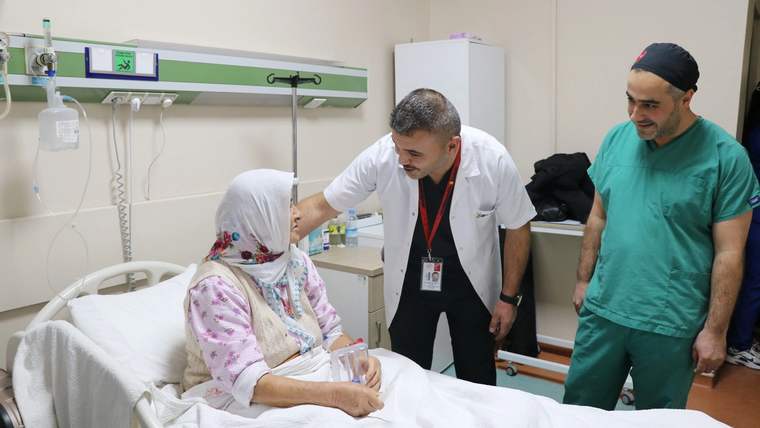 Hastane odasında yatakta oturan başörtülü bir hastanın yanında beyaz önlüklü ve yeşil formalı iki sağlık çalışanı duruyor.